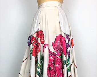 May include: A white full skirt with a floral print featuring large pink, red, and purple flowers with green leaves and gold accents. The skirt has a high waist and two large pockets on the sides.