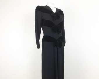May include: A long black dress with a fitted bodice and a flowing skirt. The dress features three black velvet bows on the bodice.