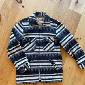 Könnte beinhalten: Eine schwarz-weiß gestreifte Jacke mit geometrischem Muster. Die Jacke hat einen Reißverschluss und zwei Pattentaschen. Das Etikett auf der Jacke lautet "Mackinaw Woolen Mills".
