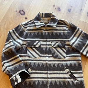Könnte beinhalten: Eine braune und weiße gestreifte Jacke mit geometrischem Muster. Die Jacke hat einen Reißverschluss und zwei Pattentaschen. Das Etikett auf der Jacke lautet "Mackinaw".