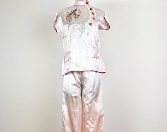 May include: A light pink satin pajama set. The top has short sleeves, a mandarin collar, and red trim. The top features an embroidered design with colorful birds and flowers. The matching pants are wide-legged.