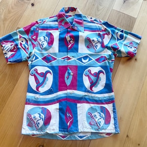 May include: A short-sleeved button-up shirt with a colorful geometric pattern. The pattern features blue, pink, and white shapes with stylized figures.