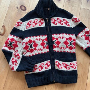 May include: A black, red, and cream-colored zip-up sweater with a shawl collar. The sweater features a geometric pattern in red and cream against a black background. It has two front pockets and ribbed cuffs.
