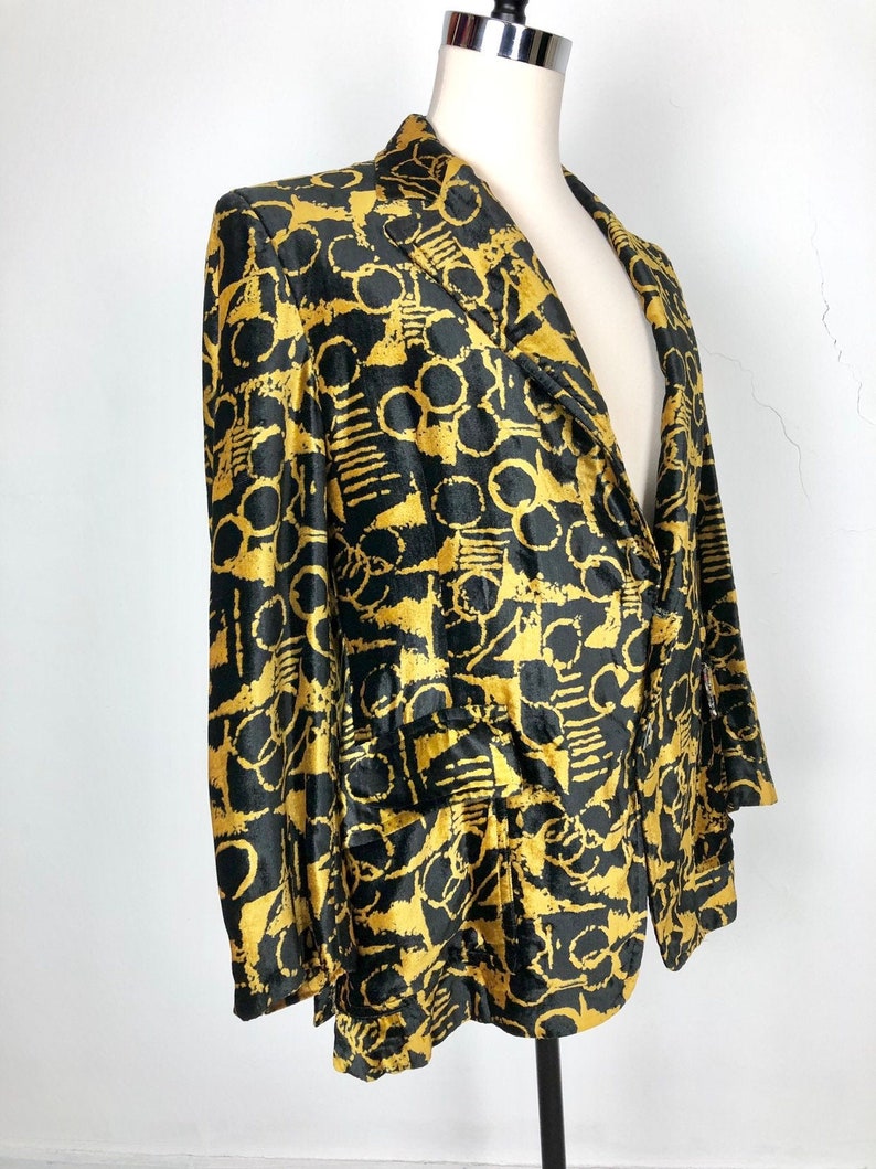 May include: A black and gold patterned blazer with a notched collar and long sleeves. The blazer has a geometric pattern with circles and lines.