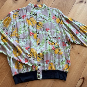 May include: A button-up shirt with a tropical banana and leaf pattern in yellow, green, and red on a light tan background. The shirt has two chest pockets, long sleeves, and a black band at the waist.