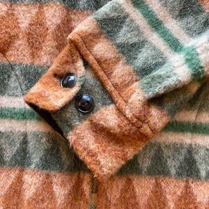 Può includere: Primo piano di una giacca a righe marroni e verdi con chiusura a zip. La giacca ha una texture soffice e due bottoni sul polsino.