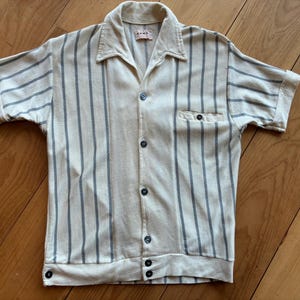May include: Cream-colored short-sleeved shirt with vertical blue-gray stripes. The shirt has a collar, a chest pocket, and a button-up front with dark buttons. The sleeves are cuffed.