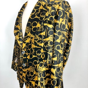 May include: A black and gold velvet blazer with a geometric pattern. The blazer has a notched collar, long sleeves, and a single-breasted closure.