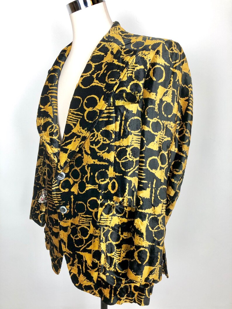 May include: A black and gold velvet blazer with a geometric pattern. The blazer has a notched collar, long sleeves, and a single-breasted closure.