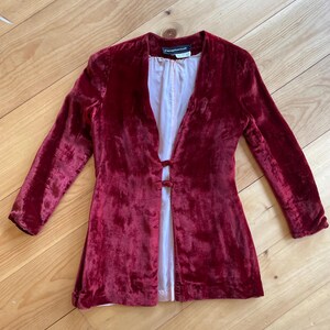 Könnte beinhalten: Burgunderfarbene Samtjacke mit langen Ärmeln und Knopfverschluss. Die Jacke hat ein weißes Futter und ein Etikett am Ausschnitt. Die Jacke liegt flach auf einer Holzoberfläche.