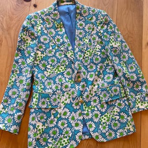 May include: A floral blazer featuring a daisy pattern in white, green, and teal. The jacket has gold buttons and a light blue lining. The blazer has two front pockets and a classic lapel design.