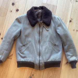 Könnte beinhalten: Eine hellgraue Bomberjacke mit einem braunen Kunstfellkragen und einem Reißverschluss. Die Jacke hat zwei Taschen auf der Vorderseite und einen gerippten Strickbund und -ärmel.