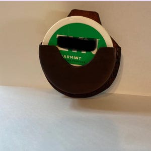 Pode incluir: Um porta-sachês de menta em couro marrom com um design circular. O suporte foi projetado para conter um recipiente redondo de menta, com a marca "ARMINT" visível no recipiente de menta.