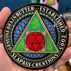 May include: A black and silver circular sticker with a green, red, and yellow geometric design. The text "Genuine Brain Butter Established 2005 Slapass Creations" is written around the edge of the sticker.
