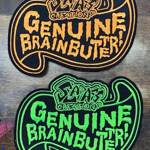 May include: Two embroidered patches with the text "Sada's Creations Genuine Brainbutter!" in orange and green. The patches are black with orange and green outlines, and the text is in a stylized font.