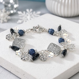 May include: A silver-toned charm bracelet featuring blue speckled round beads, black oval beads, clear faceted crystal beads, and intricate silver snowflake charms. The bracelet is secured with an ornate silver toggle clasp and square metal links.