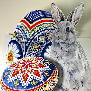May include: A 3D pop-up card featuring a gray rabbit with long ears and a blue and white patterned Easter egg. The egg has a red and yellow geometric design.