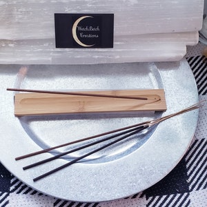 May include: A wooden incense holder with a single incense stick burning on a silver plate. The holder is sitting on top of a stack of selenite crystals. The text "Witch & Bitch Creations" is visible on a business card.
