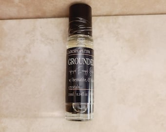 May include: A clear glass roller bottle with a black cap and a black label featuring the text WBCSHOPS.COM, GROUNDED, Infused Energy Roll, w/ hematite, & black crystals, and 10ml / 0.34fl oz. The bottle contains a transparent liquid and small dark stones.