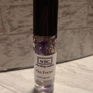 May include: A clear glass roller bottle with a black cap and purple flowers inside. The label reads "WBC wbcshop.online Fkn Focus! shake gently".