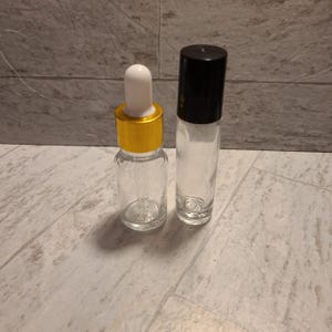 May include: Two empty glass bottles, one with a gold lid and a dropper, and the other with a black lid and a roller ball.