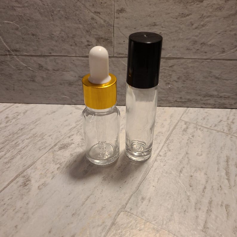 May include: Two empty glass bottles, one with a gold lid and a dropper, and the other with a black lid and a roller ball.