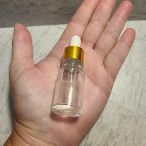 May include: A clear glass bottle with a gold lid and a white dropper. The bottle is empty and is sitting on a person's hand.