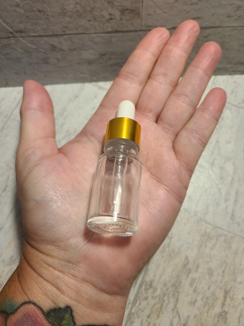 May include: A clear glass bottle with a gold lid and a white dropper. The bottle is empty and is sitting on a person's hand.