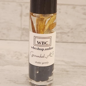 May include: A clear glass roller bottle with a black cap and a white label. The label says "WBC" and "wbc shop.online" and "Grounded AJ" and "shake gently". The bottle contains a clear liquid with dried plant material inside.