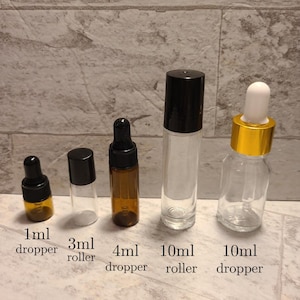 May include: Five glass bottles with different sizes and styles. The bottles are clear glass with black, gold, and brown caps. The bottles are labeled with the following sizes: 1 ml dropper, 3 ml roller, 4 ml dropper, 10 ml roller, and 10 ml dropper.