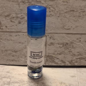 May include: A clear glass roller bottle with a bright blue cap. The bottle has a white label with the text "WBC", "wbshop.online", "Sunkissed", and "shake gently". The bottle contains small blue and green stones.