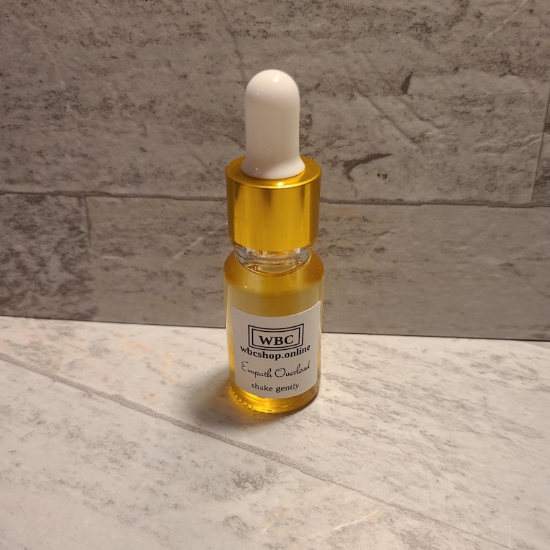 May include: A small, clear glass bottle with a gold cap and dropper. The bottle contains a yellow liquid and has a white label with the text "WBC wbcshop.online Empath Overload shake gently".