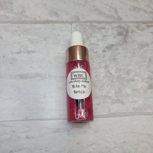 May include: A clear glass bottle with a rose-colored liquid and a gold dropper top. The bottle has a white label with the text "WBC wbc shop.online Bite Me Betch".