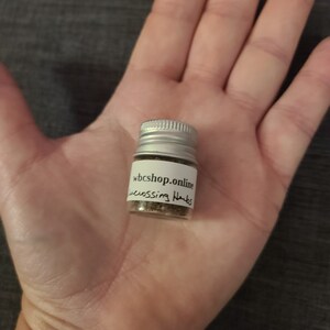 Puede incluir: Una peque&ntilde;a botella plateada con una etiqueta blanca que dice "wbshop.online" y "crossing herbs". La botella est&aacute; llena de hierbas marrones.