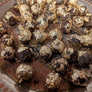 May include: A collection of dark, round food items, each partially or fully covered in gold leaf, arranged on a decorative silver platter. The gold leaf adds a luxurious, shimmering effect to the treats.