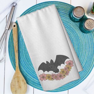 May include: White waffle-weave kitchen towel featuring a gray bat design surrounded by pink and yellow flowers. The towel is folded and placed on a blue and green placemat, with a wooden spoon and whisk visible.