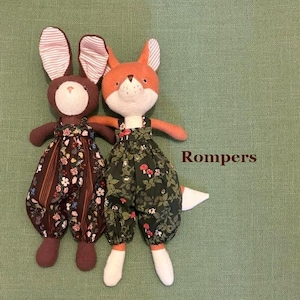 May include: Two plush toys, a brown rabbit and an orange fox, both wearing fabric rompers. The rabbit wears a dark brown romper with a floral and striped pattern. The fox wears a dark green romper with a mushroom and leaf print. Text reads Rompers.