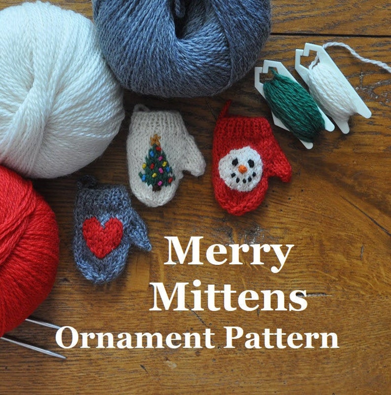 Four knitted mitten ornaments with Christmas tree, snowman, and heart designs on a wooden surface with yarn.