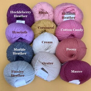 Puede incluir: Una colecci&oacute;n de ovillos de lana en varios colores, incluyendo morado, rosa, amarillo y gris. Cada ovillo est&aacute; etiquetado con su nombre de color, como "Huckleberry Heather", "Blush" y "Finnley Heather". La lana est&aacute; dispuesta sobre un fondo amarillo.