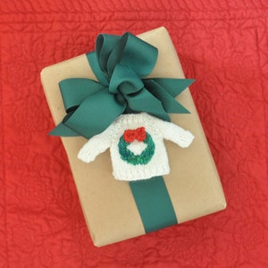 May include: A brown gift box with a green ribbon and bow. A small knitted sweater with a green wreath design is attached to the bow.