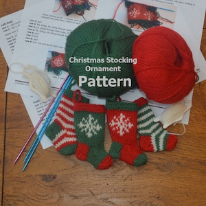 Peut inclure: Quatre ornements de chaussettes de Noël en tricot, deux avec des rayures rouges et blanches, un avec un motif de flocon de neige vert et blanc et un avec un motif de flocon de neige rouge et blanc. Les ornements sont sur une surface en bois avec des pelotes de laine et des aiguilles à tricoter.