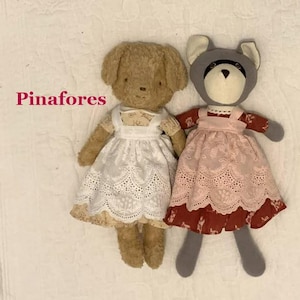Puede incluir: Dos muñecos de peluche con el texto Pinafores. El muñeco de la izquierda es de color canela con un delantal blanco de ojales sobre un vestido estampado. El de la derecha es un mapache gris con un delantal rosa sobre un vestido rojo oscuro.