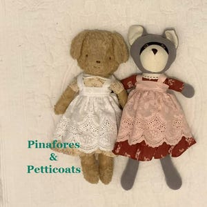 Peut inclure: Deux poupées en peluche, une figure d'ours beige et une figure de raton laveur gris. L'ours porte un tablier blanc à œillets sur une robe à motifs. Le raton laveur porte un tablier rose à œillets sur une robe rouge foncé à motifs. Texte : Pinafores & Petticoats.