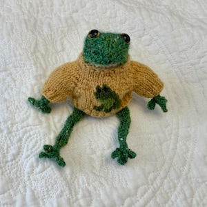 May include: Hand-knitted frog plushie with a green head and legs, wearing a tan sweater with a green frog applique. The frog has black button eyes and is posed on a white quilted surface. A whimsical, handmade item.