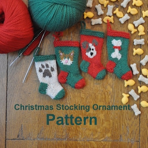 Puede incluir: Cuatro pequeñas medias de Navidad de punto con diferentes diseños: una huella de pata, una cara de gato, una cara de perro y un hueso. Las medias son rojas, verdes y blancas y están sobre una superficie de madera con una bola de lana verde y una bola de lana roja.