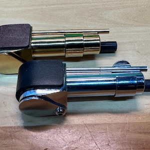 以下が含まれることがあります： 革のアクセントが付いた2つの金属製葉巻ライター。1つのライターは茶色の革のアクセントと金色の金属です。もう1つのライターは黒い革のアクセントと銀色の金属です。どちらのライターにも黒いノズルが付いています。
