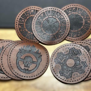 K&ouml;nnte beinhalten: Satz von zehn braunen Lederuntersetzern mit verschiedenen schwarzen Designs. Die Designs beinhalten einen Sch&auml;del und Flammen, ein Zahnrad und einen Kreis mit Symbolen.