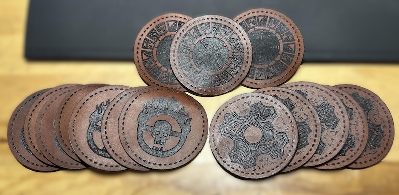 K&ouml;nnte beinhalten: Satz von zehn braunen Lederuntersetzern mit verschiedenen schwarzen Designs. Die Designs beinhalten einen Sch&auml;del und Flammen, ein Zahnrad und einen Kreis mit Symbolen.