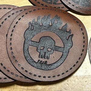 May include: A brown leather coaster with a black stitched border and a black and white skull with flames design. The design is a stylized skull with flames around it and a circle with a line through it.