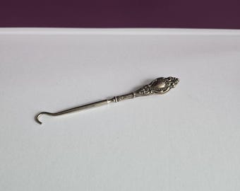 May include: A silver-toned metal button hook featuring an ornate, embossed handle with intricate scrollwork and floral detailing. The tool has a long, slender shaft that terminates in a small, curved hook designed for fastening buttons on clothing.